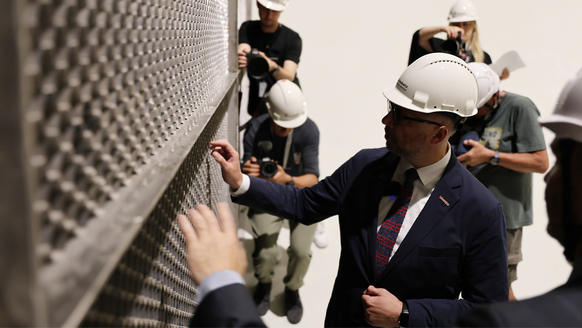Minister Kulasek dotyka metalowej ściany z tunelami wyprostowującymi rozpędzone powietrze, tuż przed wejściem do przestrzeni badawczej | źródło: Łukasiewicz - ILOT